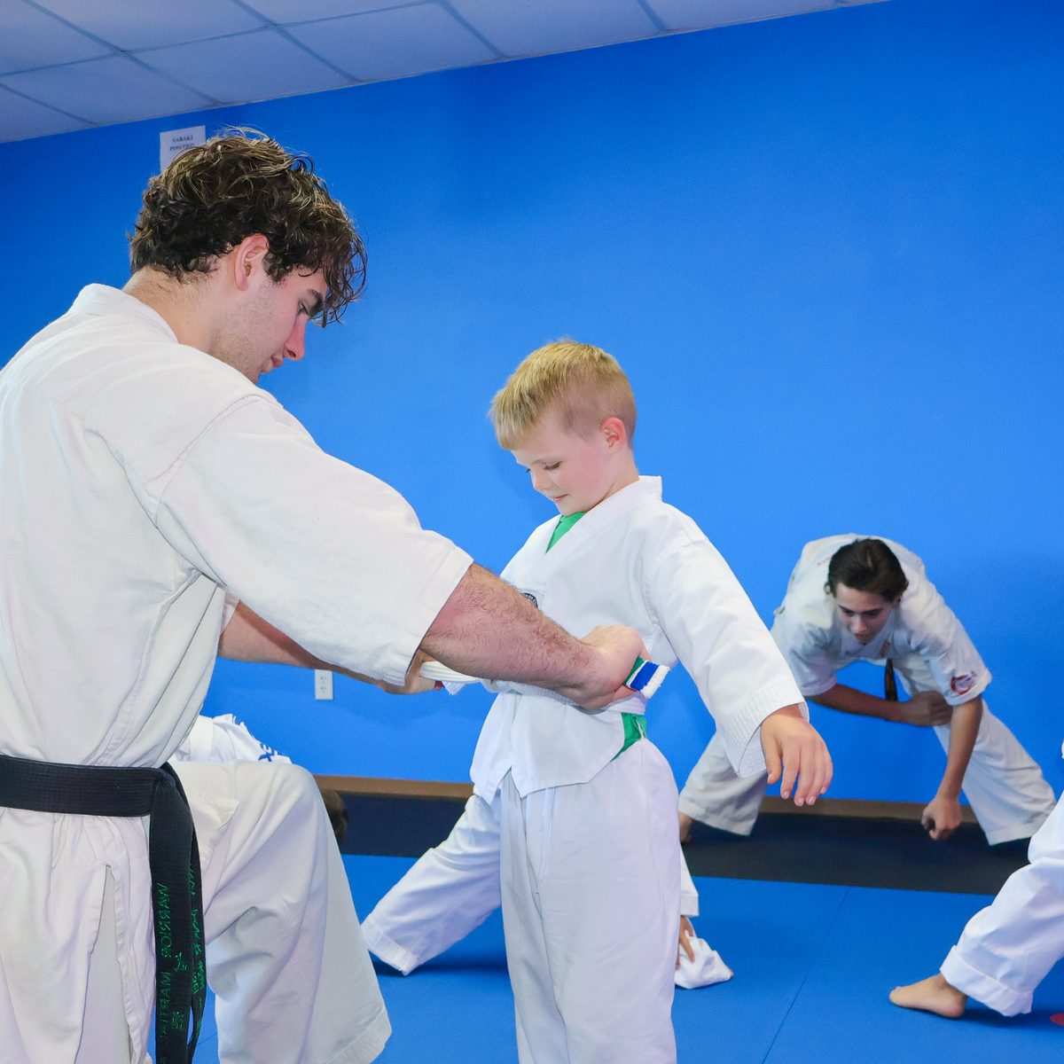 A black-belt instructor helping a young student tie his green belt while other students train on the mats behind them